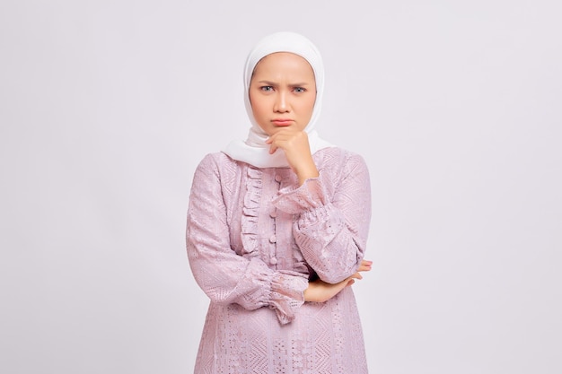 Porträt einer nachdenklichen, schönen jungen asiatischen muslimischen Frau mit Hijab und lila Kleid, die ihr Kinn berührt und isoliert auf weißem Studiohintergrund in die Kamera blickt