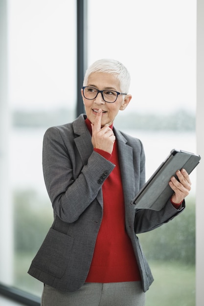 Porträt einer nachdenklichen Senior-Geschäftsfrau, die an einem digitalen Tablet arbeitet, während sie in einem modernen Büro in der Nähe des Fensters steht.