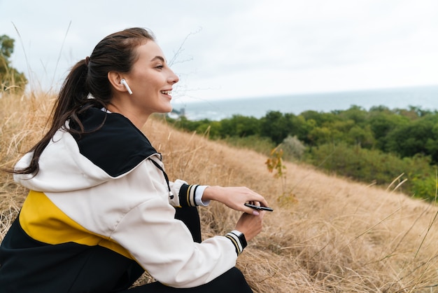 Porträt einer netten, glücklichen Frau in Sportkleidung mit Ohrstöpseln und Smartphone, während sie auf trockenem Gras am Meer sitzt
