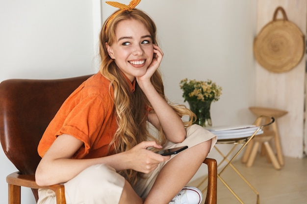Porträt einer netten glücklichen lächelnden jungen blonden Frau zu Hause zuhause unter Verwendung des Handys, das auf Stuhl sitzt.