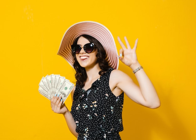 Porträt einer netten jungen Frau mit der Sonnenbrille und Hut, die Geldbanknoten halten und über gelber Wand lokalisiert feiern