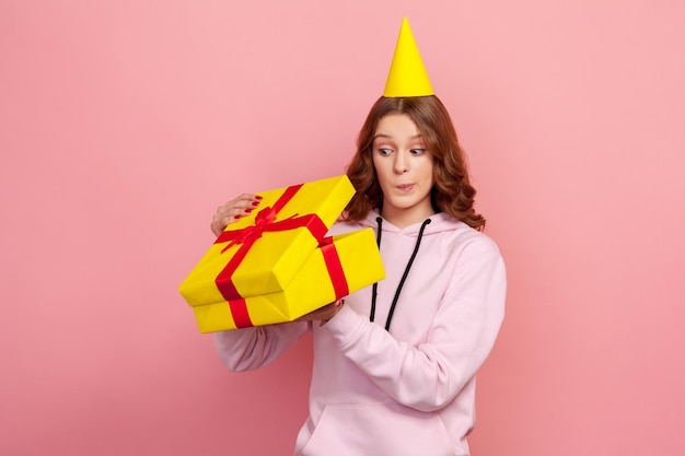 Porträt einer neugierigen Teenager-Frau in Hoodie mit Partyhut, die heimlich in ein verpacktes Geschenkbox-Geburtstagsgeschenk lugt Studioaufnahme isoliert auf rosa Hintergrund
