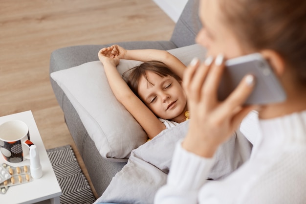 Porträt einer nicht erkennbaren Mutter, die wegen der Krankheit ihrer Tochter einen Arzt anruft, das Kind liegt auf dem Sofa auf dem Kissen unter der Decke, leidet unter Kopfschmerzen und hoher Temperatur.