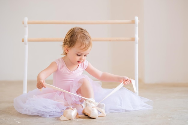 Porträt einer niedlichen blauäugigen Babyballerina in Rosa, die in der Nähe der Ballettstange sitzt und Spitzenschuhe anzieht