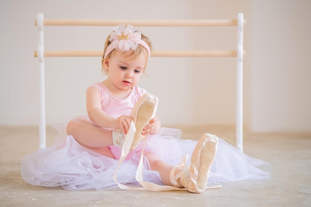 Porträt einer niedlichen blauäugigen Babyballerina in Rosa, die in der Nähe der Ballettstange sitzt und Spitzenschuhe anzieht