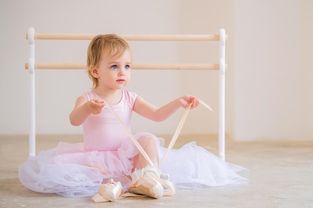 Porträt einer niedlichen blauäugigen Babyballerina in Rosa, die in der Nähe der Ballettstange sitzt und Spitzenschuhe anzieht