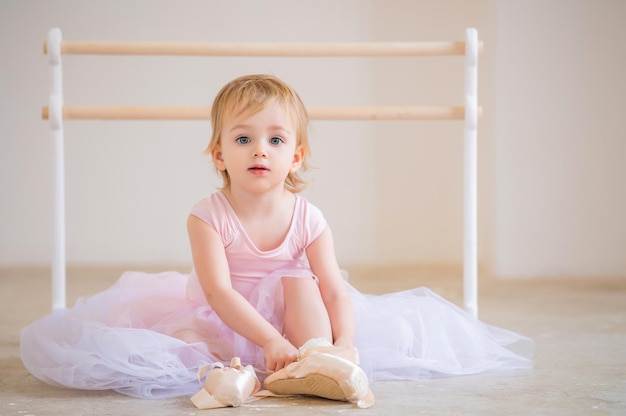 Porträt einer niedlichen blauäugigen Babyballerina in Rosa, die in der Nähe der Ballettstange sitzt und Spitzenschuhe anzieht