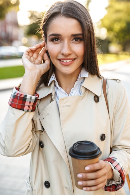 Porträt einer optimistischen, süßen Frau mit Mantel, die Kaffee zum Mitnehmen trinkt und beim Gehen auf der Stadtstraße lächelt