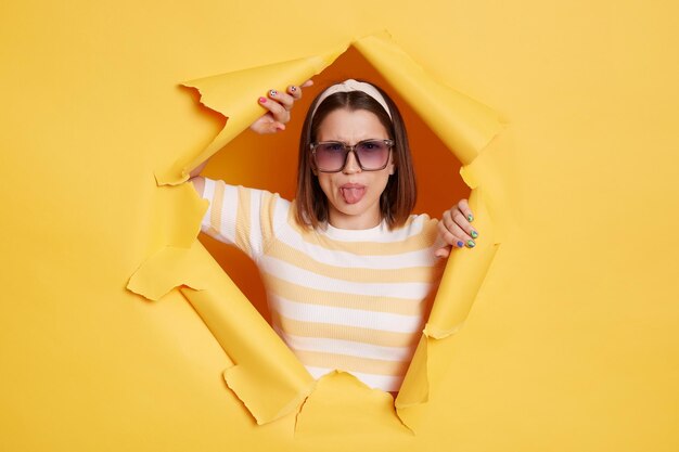Porträt einer positiv optimistischen Frau mit gestreiftem T-Shirt-Haarband und Sonnenbrille, die durch ein Papierloch in einer gelben Wand bricht und in die Kamera blickt, die die Zunge herausstreckt