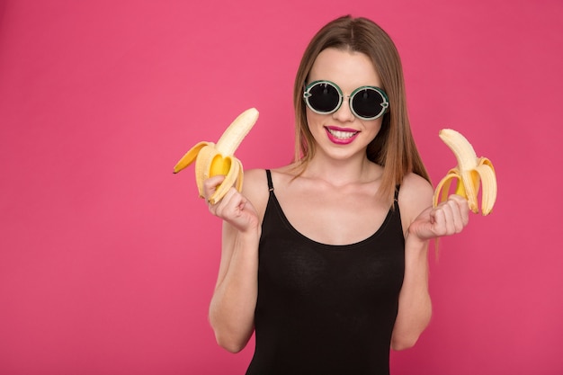 Porträt einer positiven, attraktiven, fröhlichen, fröhlichen jungen Frau mit schwarzer Sonnenbrille, die zwei Bananen an rosa Wand hält