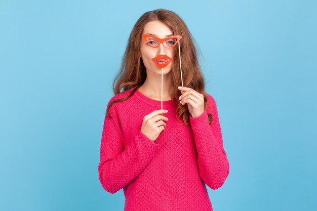 Porträt einer positiven Frau in rosafarbenem Pullover, die Papierbrille und Lippen hält, in die Kamera schaut, festliche Stimmung hat, Party-Requisiten trägt. Studioaufnahme im Innenbereich isoliert auf blauem Hintergrund.