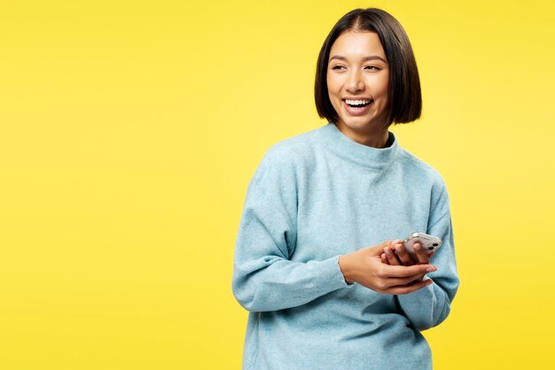 Foto porträt einer positiven schönen asiatischen frau, die einen beiläufigen pullover trägt, ein handy hält und im gespräch ist, die auf den kopierraum wegblickt, die auf einem gelben hintergrund isoliert steht.