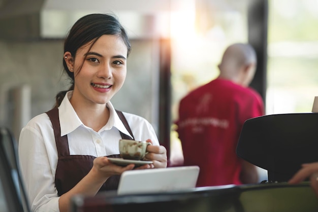 Porträt einer professionellen asiatischen Barista-Frau in Schürze, die eine Tasse heißen Kaffee im Café hält