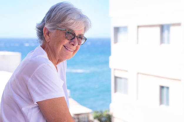 Porträt einer reifen attraktiven Frau, die sich auf ihrem Hausbalkon entspannt und das Meer und den blauen Himmel an einem sonnigen Tag im Freien betrachtet.