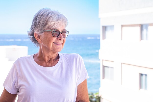 Porträt einer reifen attraktiven grauhaarigen Frau, die sich an einem sonnigen Tag im Freien auf ihrem Hausbalkon, dem Hintergrund des Meeres und des blauen Himmels entspannt.