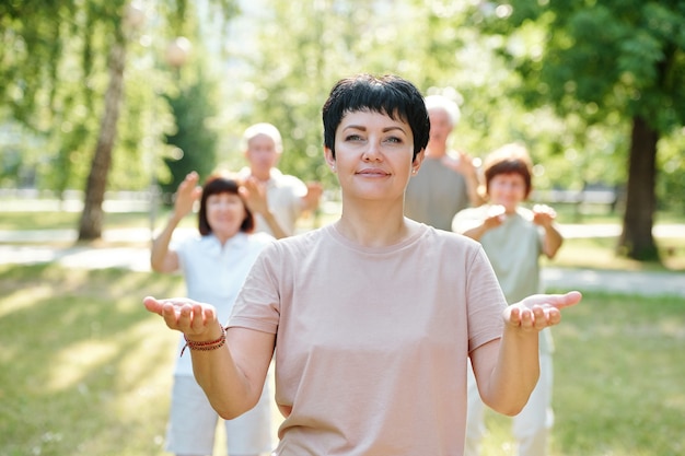 Porträt einer reifen Frau, die in die Kamera schaut und zusammen mit anderen Leuten im Hintergrund Yoga macht