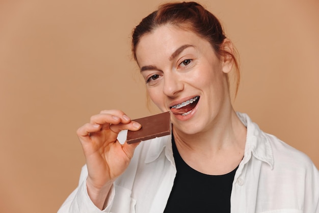 Porträt einer reifen Frau in einer Zahnspange, die Schokolade auf einem beigen Hintergrund beißt