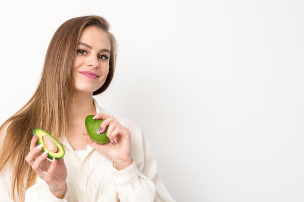 Porträt einer reizend lächelnden jungen brünetten kaukasischen Frau, die das weiße Hemd mit dem langen Haar trägt und die Avocadostellung lokalisiert über weißem Hintergrund hält und zeigt