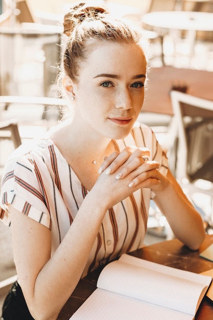 Porträt einer reizenden jungen Frau mit roten Haaren und Sommersprossen, die Kamera lächelnd betrachten. Junge niedliche Schriftstellerin, die an ihrem Buch draußen arbeitet.