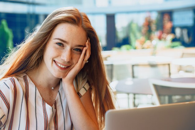 Porträt einer reizenden jungen Frau mit roten Haaren und Sommersprossen lächelnd, während Gesicht mit einer Hand berührend, die an einem Laptop im Freien in einem Café arbeitet.