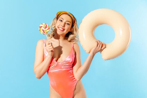 Porträt einer reizenden, sorglosen, schönen Frau im Badeanzug, die einen Gummiring und süße Lollipop-Süßigkeiten hält und sich am tropischen Strand, Ferienort, ausruht. Studioaufnahme im Innenbereich isoliert auf blauem Hintergrund