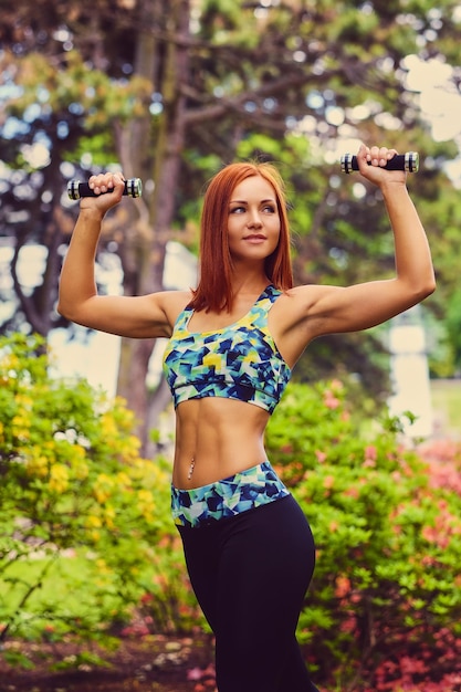 Porträt einer rothaarigen Fitness-Frau hält Hanteln in einem Park im Freien.