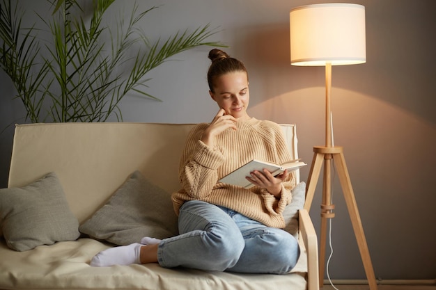 Porträt einer ruhigen lächelnden Frau mit braunen Haaren, die einen beigen Pullover und Jeans trägt und auf Husten sitzt, liest ein Buch, das sich auf ein Seminar vorbereitet oder Literatur liest
