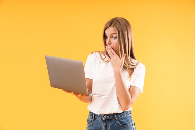 Porträt einer schockierten blonden Frau, die ein lässiges T-Shirt trägt, das Wunder ausdrückt, während sie den Laptop isoliert über der gelben Wand im Studio benutzt