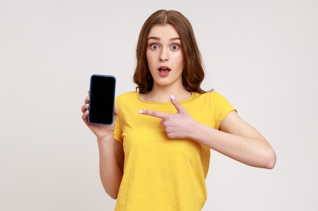 Porträt einer schockierten braunhaarigen Frau mit welligem Haar in gelbem T-Shirt, die auf das Handy zeigt und mit Erstaunen und großen Augen in die Kamera schaut Studioaufnahme isoliert auf grauem Hintergrund