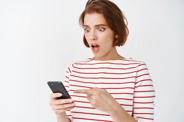 Porträt einer schockierten Frau, die mit besorgtem und verwirrtem Gesicht auf das Smartphone schaut und schlechte Nachrichten auf dem mobilen Bildschirm liest, der vor weißem Hintergrund steht
