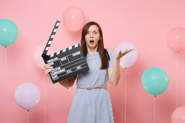 Porträt einer schockierten Frau in blauem Kleid, die die Hände ausbreitet, die klassische schwarze Filmklappe auf rosafarbenem Hintergrund mit bunten Luftballons halten. Geburtstagsfeier, Menschen aufrichtige Emotionen.