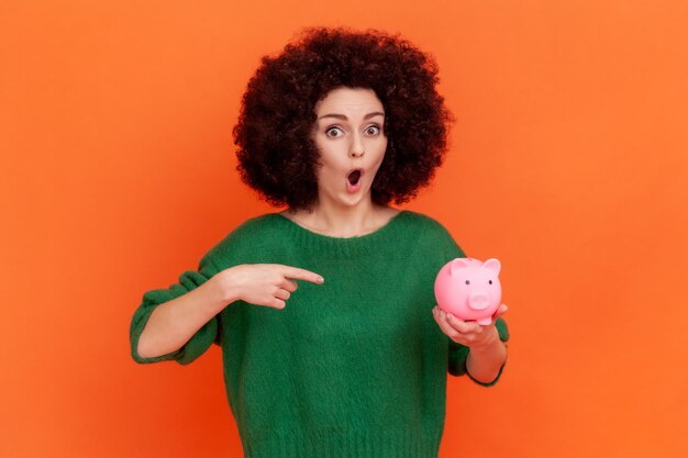 Porträt einer schockierten Frau mit Afro-Frisur, die einen grünen Pullover im lässigen Stil trägt, der auf das Sparschwein in ihrer Hand zeigt vorteilhaftes Bankangebot Studioaufnahme im Innenbereich isoliert auf orangefarbenem Hintergrund