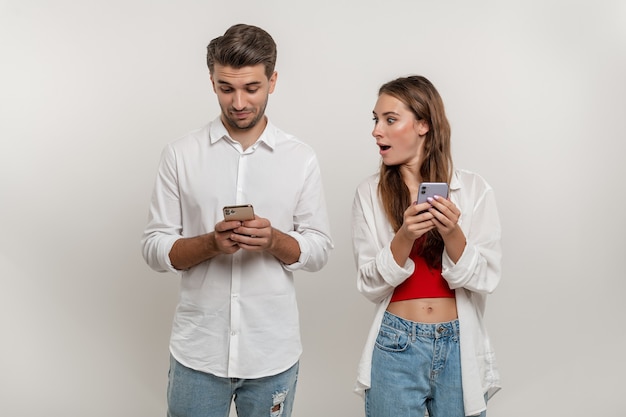 Porträt einer schockierten kaukasischen Frau, die ihren Freund mit Handy auf weißem Hintergrund ausspioniert