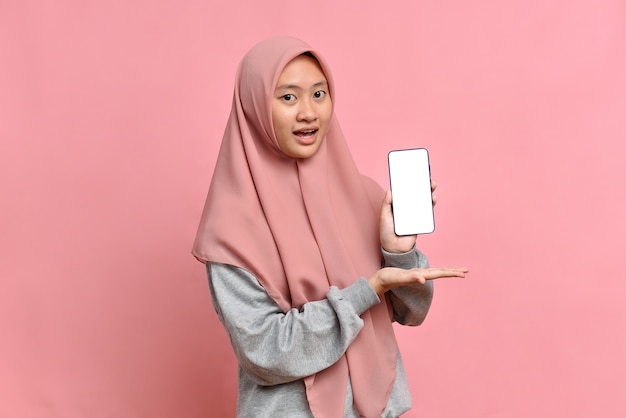 Porträt einer schockierten schönen asiatischen muslimischen Frau, die weißen Bildschirm am Telefon auf rosafarbenem Hintergrund zeigt
