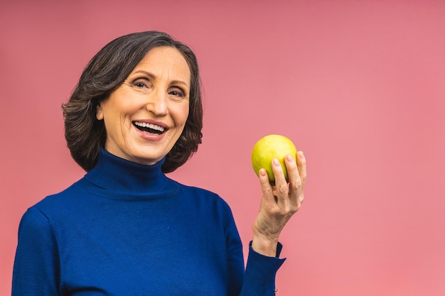 Porträt einer schönen älteren reifen gealterten Frau, die einen Apfel hält, lächelnd, lokalisiert über rosa Hintergrund. Ein Apfel am Tag hält einen Arzt fern.