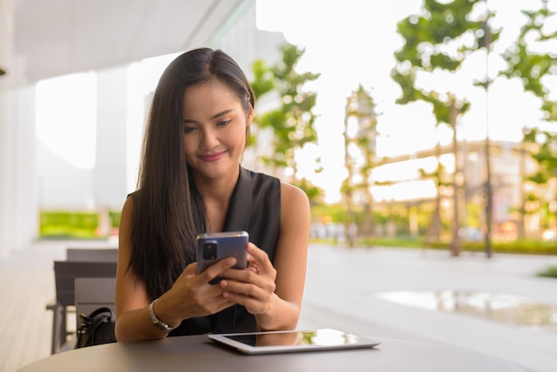 Porträt einer schönen asiatischen Frau, die draußen im Café-Restaurant mit Handy sitzt