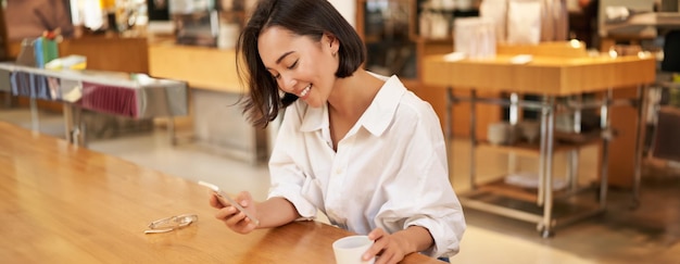 Porträt einer schönen asiatischen Frau mit Smartphone, die sich im Café entspannt und dabei Kaffee genießt