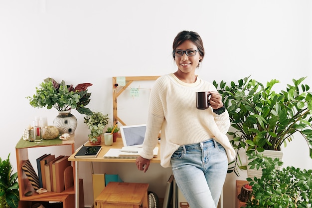 Porträt einer schönen attraktiven jungen Grafikdesignerin, die an ihrem Tisch im Home Office steht, eine große Tasse Kaffee trinkt und wegschaut