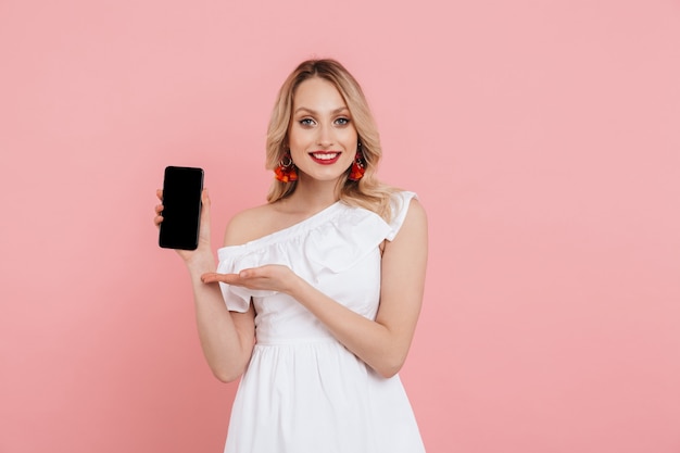 Porträt einer schönen blonden Frau, die ein Sommerkleid trägt, das isoliert über rosa steht und ein leeres Bildschirm-Handy zeigt