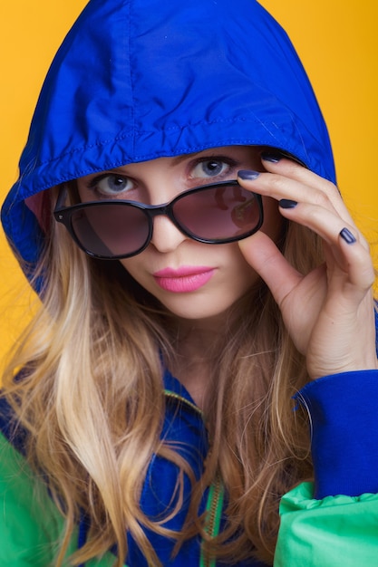 Porträt einer schönen blonden Frau mit Sonnenbrille und blaugrüner Kapuzenjacke auf gelbem Hintergrund...