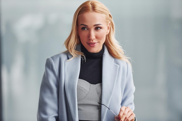 Porträt einer schönen Blondine in eleganter Kleidung, die drinnen vor grauem Hintergrund steht