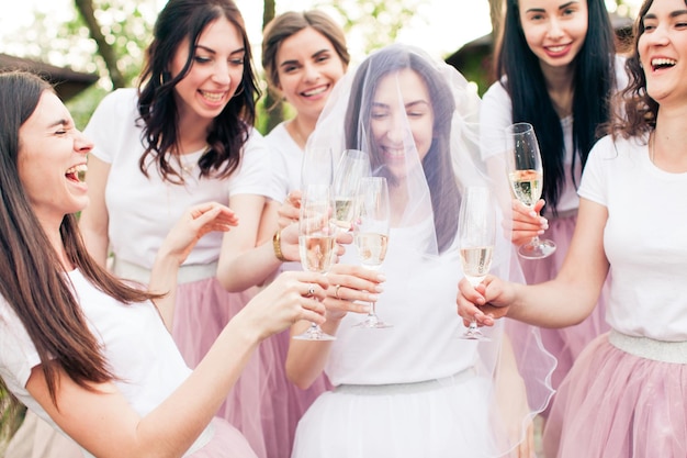 Porträt einer schönen Braut mit weißem Schleier im Gesicht, die zwischen glücklich lächelnden Brautjungfern steht, die alle Gläser mit Champagner in den Händen halten. Fröhliches Lifestyle-Konzept