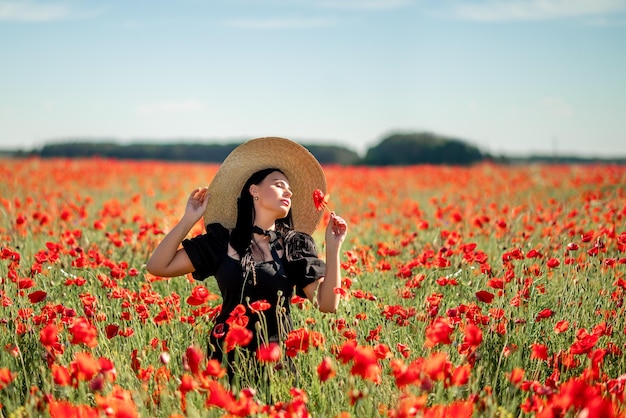 Porträt einer schönen brünetten Frau in einem Mohnfeld. Sie trägt einen großen Hut und eine rote Blume in ihren Händen