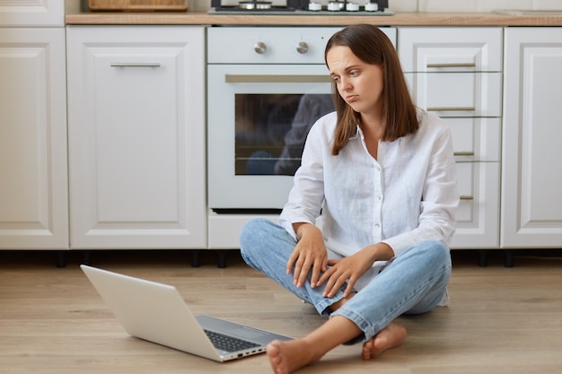 Porträt einer schönen dunkelhaarigen Frau, die auf dem Boden in der Küche sitzt, Laptop-Display mit traurigem Gesichtsausdruck betrachtet, weißes Hemd und Jeans trägt.