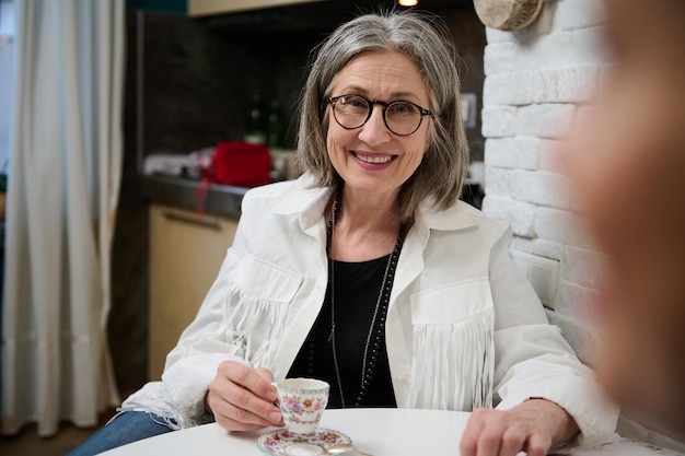 Porträt einer schönen eleganten europäischen Seniorin in stilvoller Kleidung, die am Tisch sitzt und eine Kaffeepause in ihrem gemütlichen Studio genießt