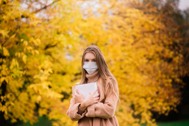 Porträt einer schönen erwachsenen jungen Frau auf dem Hintergrund des Herbstes im Park in der medizinischen Gesichtsmaske