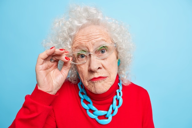 Porträt einer schönen faltigen seniorin hat lockiges graues haar, helle make-up-maniküre sieht aufmerksam durch transparente brille aus, trägt roten pullover und halskette