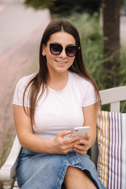 Porträt einer schönen Frau, die draußen auf einer Bank sitzt und ein Telefon benutzt. Frau mit Sonnenbrille