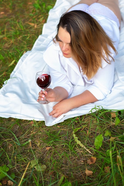 Porträt einer schönen Frau, die in einem weißen Hemd und einem durchbrochenen Höschen mit einem Glas auf dem Rasen liegt