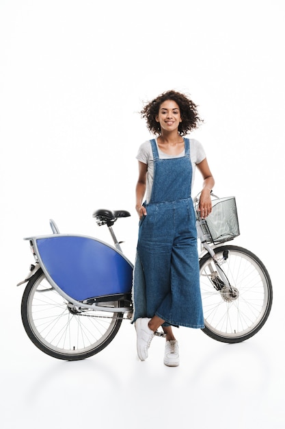 Porträt einer schönen Frau, die in Jeans-Overalls gekleidet ist und lächelt, während sie über dem Fahrrad steht, isoliert über der weißen Wand?